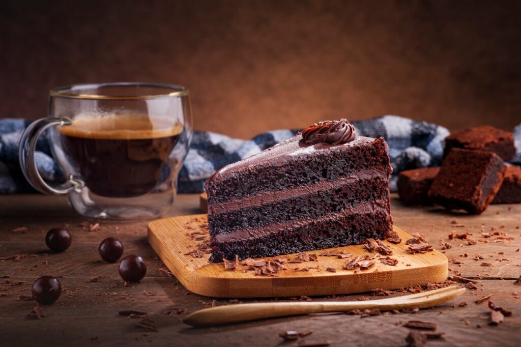 Imagem de um pedaço de bolo de chocolate com destaque na frente, e uma xícara de café ao fundo. Ambos em cima de uma mesa de madeira, e pedaços e chocolate sobre a mesa.